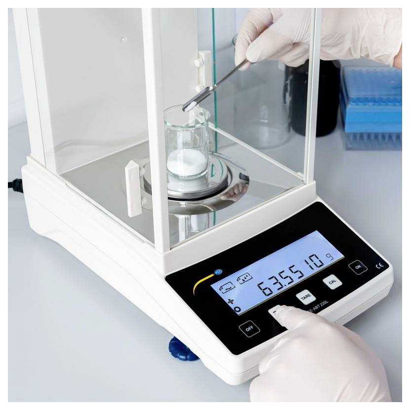 Behandschuhte Hände verwenden eine analytische Laborwaage, um eine Probe in einem Becher zu wiegen, wobei das digitale Display 6355,18 g anzeigt.