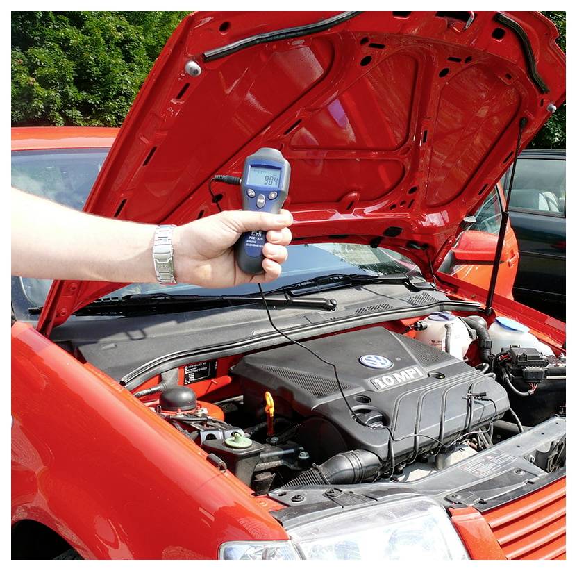 Tragbares Infrarot-Thermometer, das auf den offenen Motorraum eines roten Autos gerichtet ist und eine Temperaturanzeige während der Überprüfung der Motorwärme anzeigt.