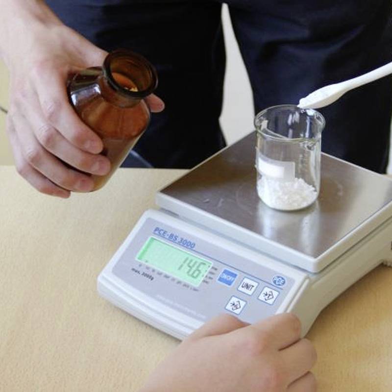 Ein Glas mit weißem Pulver wird auf einer elektronischen Waage gewogen. Eine Hand hält ein braunes Fläschchen. Gewichtsanzeige: 14,6 g.
