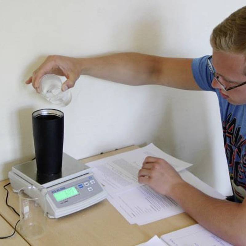 Eine Person gießt Wasser aus einem Messbecher in einen schwarzen Behälter auf einer digitalen Waage, die auf einem Tisch steht.