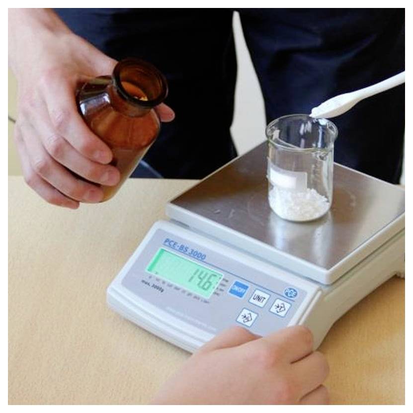 Eine Person wiegt weißes Pulver in einem Glasecher auf einer digitalen Waage ab, während sie eine braune Chemieflasche hält.
