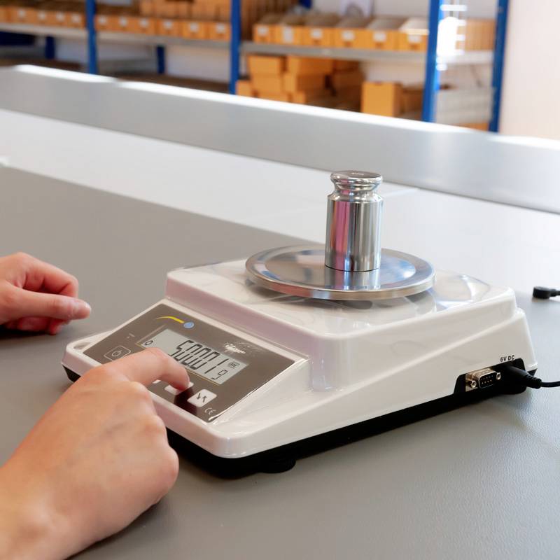 Digitale Waage mit Gewicht auf der Plattform zeigt '5000.19' Gramm an. Zwei Hände sind sichtbar; Fokus auf Messgerät.