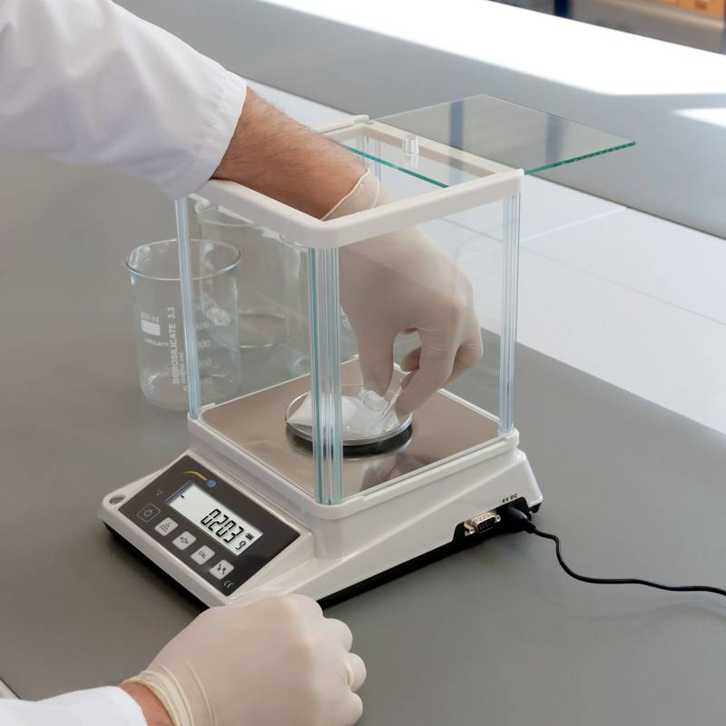 Eine Person wiegt eine Substanz auf einer digitalen Laborwaage. Neben der Waage steht ein Becherglas.