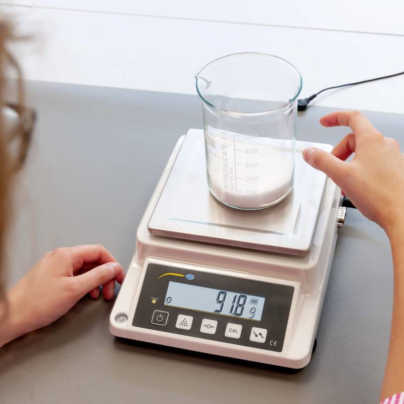 Eine Hand stellt einen Messzylinder mit Flüssigkeit auf eine digitale Waage, die ein Gewicht von 918,9 Gramm anzeigt.