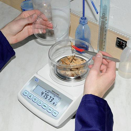 Zwei Hände legen Holzstücke in einen Glasbehälter auf einer elektronischen Waage. Das Display zeigt 45,73 g an.