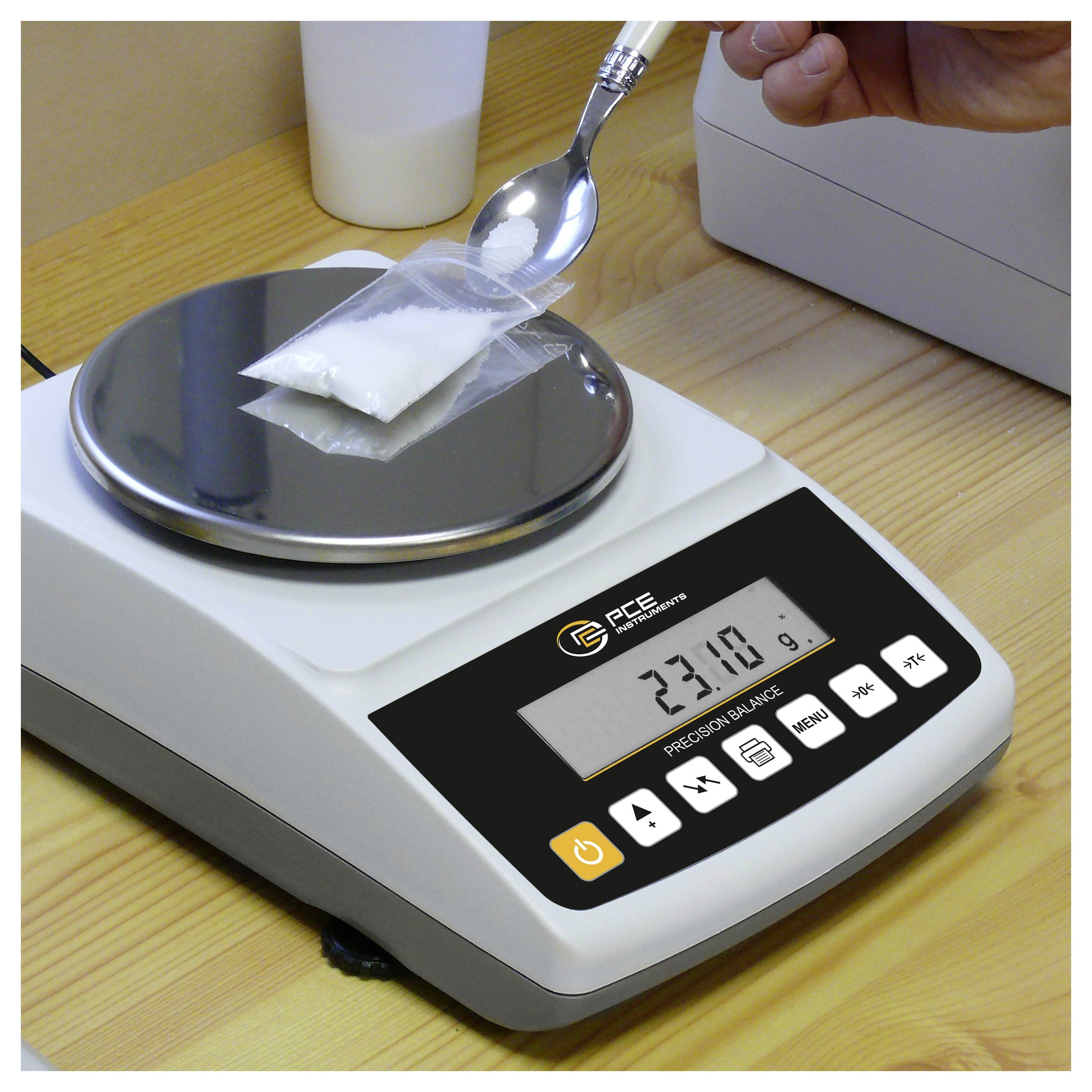 Eine digitale Waage wiegt ein kleines Kunststoffpaket, während ein Löffel Pulver hinzufügt; die Anzeige zeigt 23,0 g an.