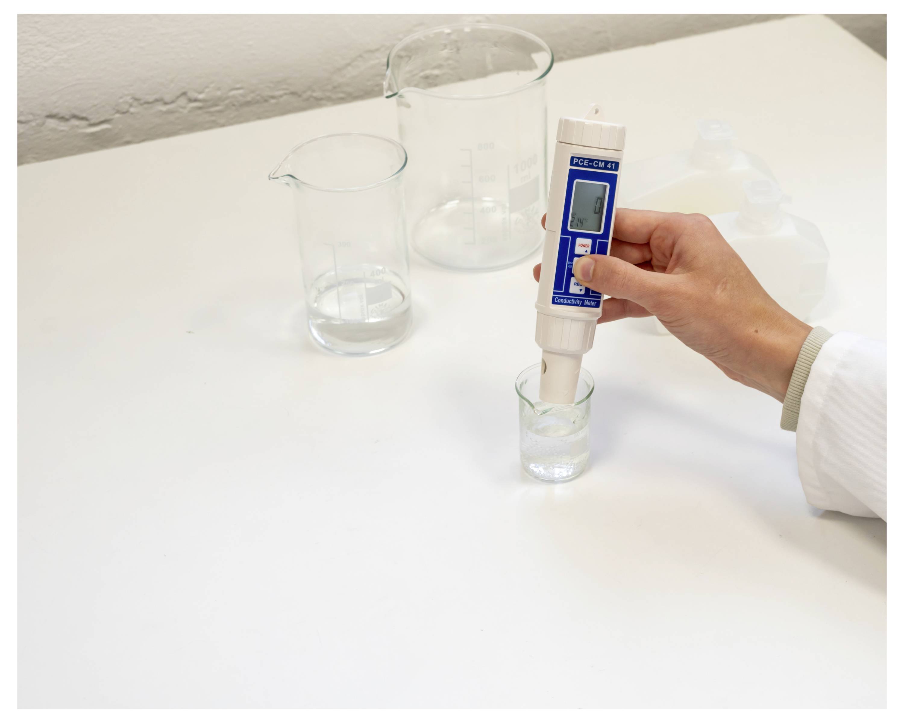 Eine Hand hält ein digitales pH-Messgerät in einem kleinen Glas mit Flüssigkeit auf einer Laborbank, mit zwei größeren Bechergläsern mit klarer Flüssigkeit im Hintergrund.