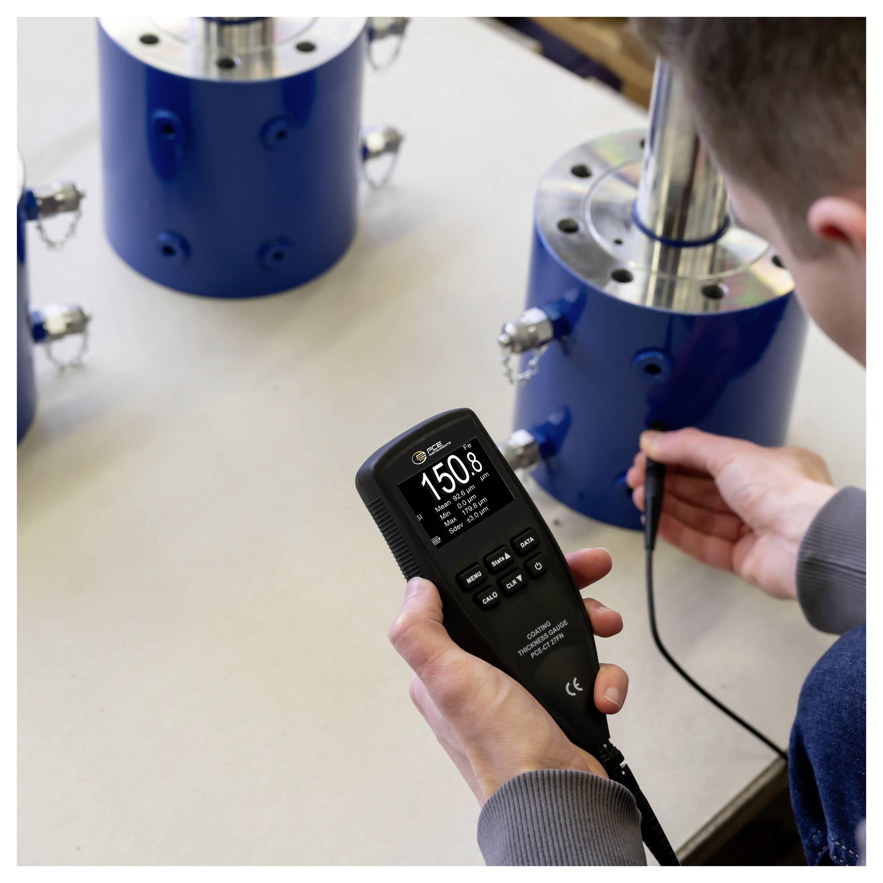 Eine Person, die ein tragbares digitales Messinstrument verwendet, um einen blauen Metallzylinder in einer Werkstatt oder einem Labor zu messen.