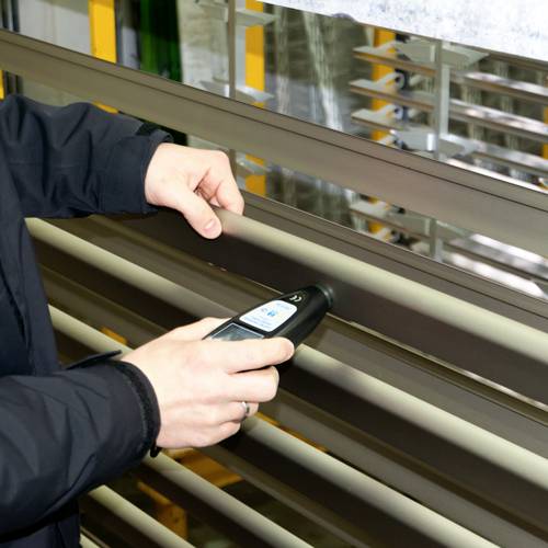 Eine Person benutzt ein Handgerät, um Jalousien zu messen oder zu justieren. Die Szene ist auf Benutzung und Funktionalität fokussiert.