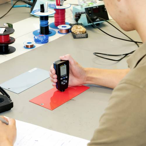 Ein Mensch misst die Dicke eines roten Materials mit einem digitalen Messgerät an einem Arbeitstisch mit Werkzeugen und Kabelrollen.