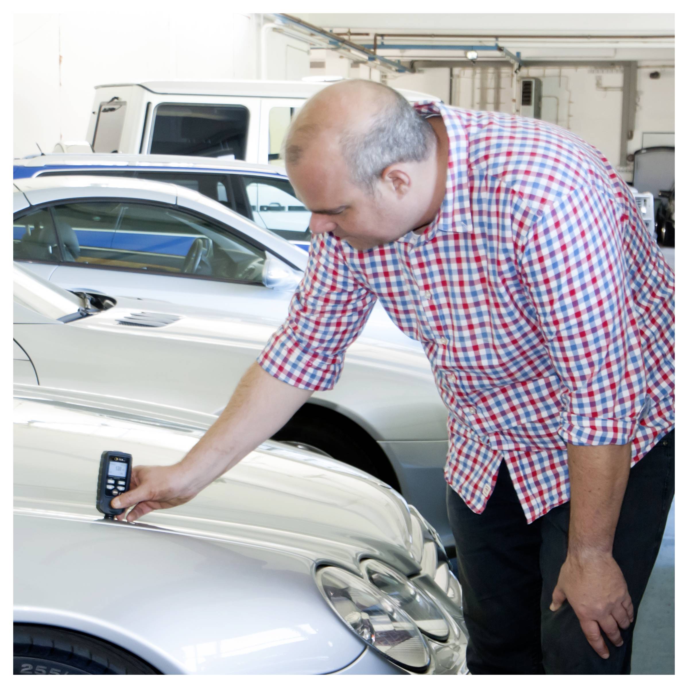 Ein Mann inspiziert die Vorderseite eines silbernen Autos in einer Garage und scheint ein tragbares Gerät auf der Motorhaube zu verwenden.