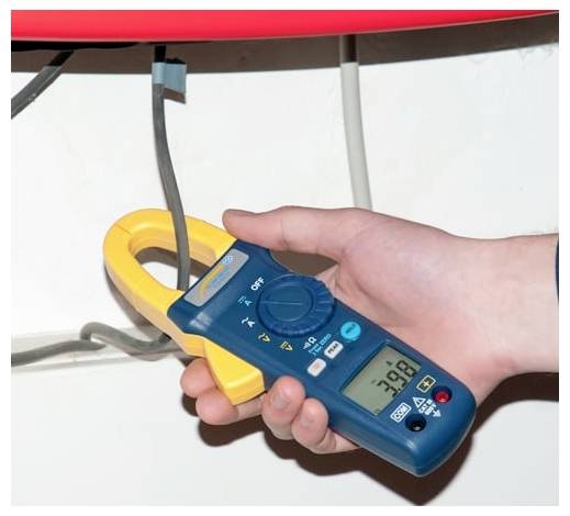 Hand hält ein Zangenmultimeter um ein Elektrokabel, wobei eine Live-Strommessung auf dem Display angezeigt wird.