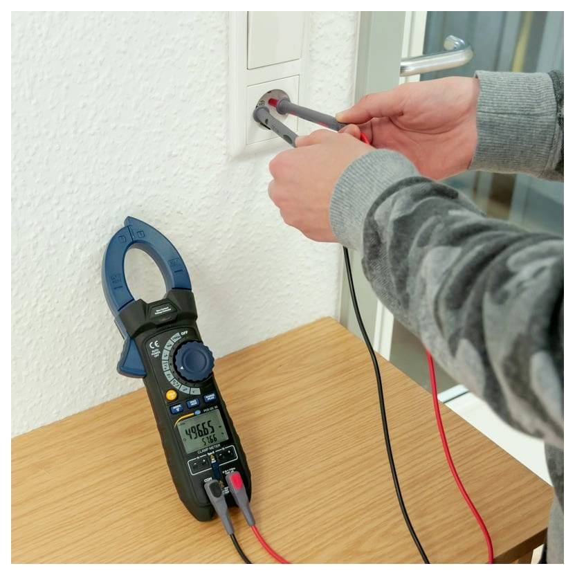 Person, die ein Stromzangenmessgerät verwendet, um eine Steckdose zu testen; das Messgerät liegt auf einem Tisch mit angebrachten roten und schwarzen Messleitungen.