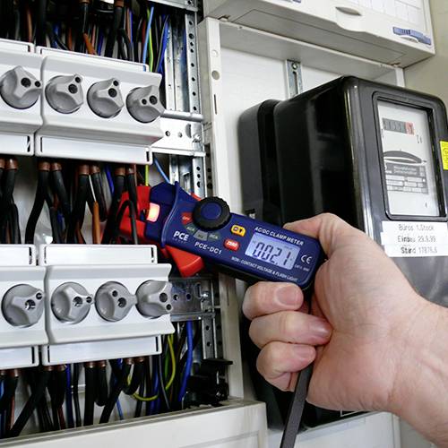 Eine Hand hält ein digitales Messgerät, das den Strom in einem elektrischen Verteilerkasten misst.
