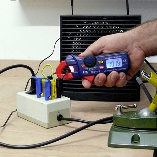 Ein Handwerker misst den Stromfluss in einem Kabel mit einer strombetriebenen Klemme. Im Hintergrund ein Verteilerkasten.