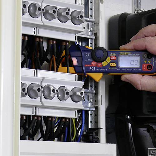 Eine Person misst die Spannung in einem Sicherungskasten mit einem Spannungsmessgerät. Kabel sind sichtbar im offenen Kasten.