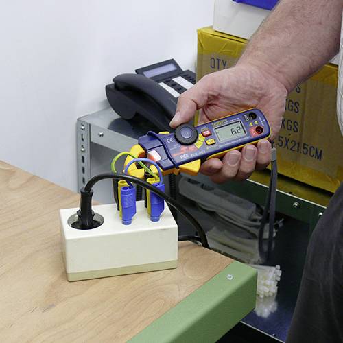 Eine Person hält ein Messgerät, das den Stromverbrauch von Elektrogeräten anzeigt. Das Display zeigt '62'.