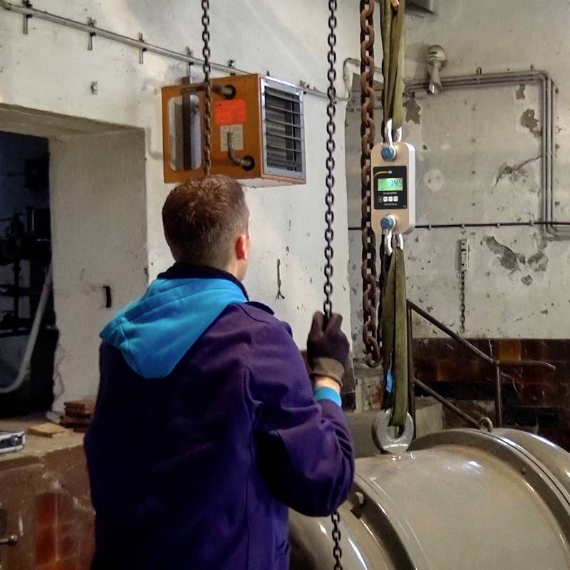 Eine Person bedient an einem industriellen Ort eine digitale Waage, die an einer Kette hängt. Die Umgebung ist funktional eingerichtet.