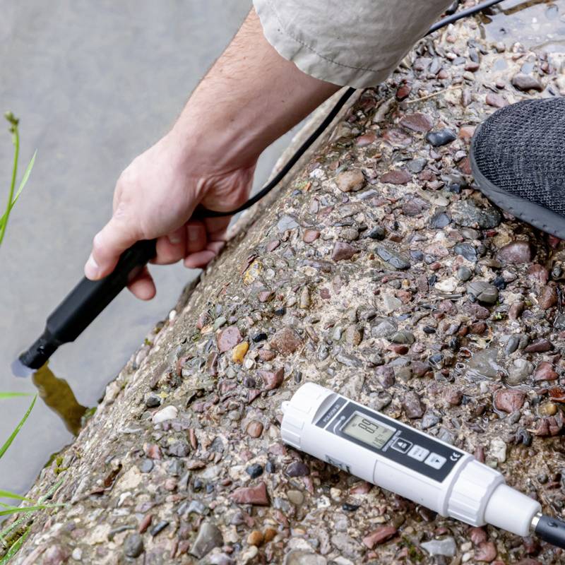 Ein Mann misst den pH-Wert des Wassers in einem Bach mit einem tragbaren, elektronischen Messgerät.