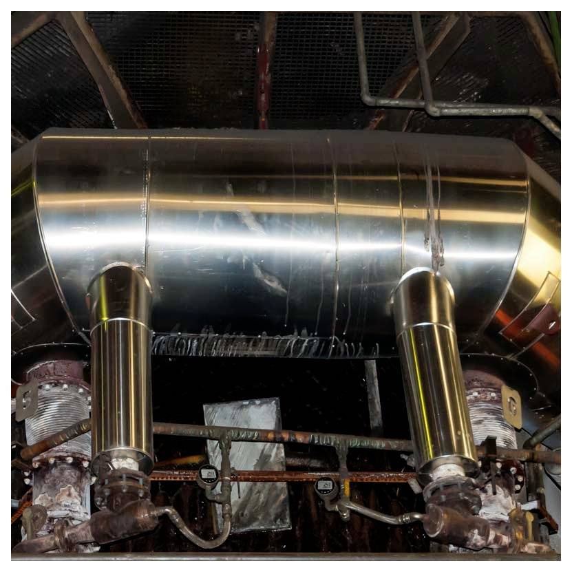 Großer polierter Metalltank oder Kanal in einem industriellen Maschinenraum, gestützt durch zwei vertikale Rohre und umgeben von Ventilen und Verkabelung.
