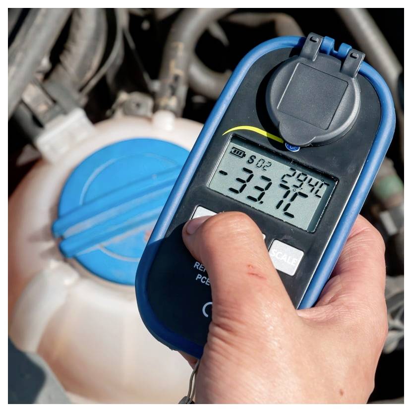 Hand hält ein Kältemittelmanometer in der Nähe eines Fahrzeugkühlmittelbehälters und zeigt eine digitale Anzeige von -33,1 °C.