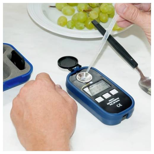 Hand, die ein tragbares Refraktometer benutzt, um eine Traubenprobe auf einer Tischplatte zu testen.