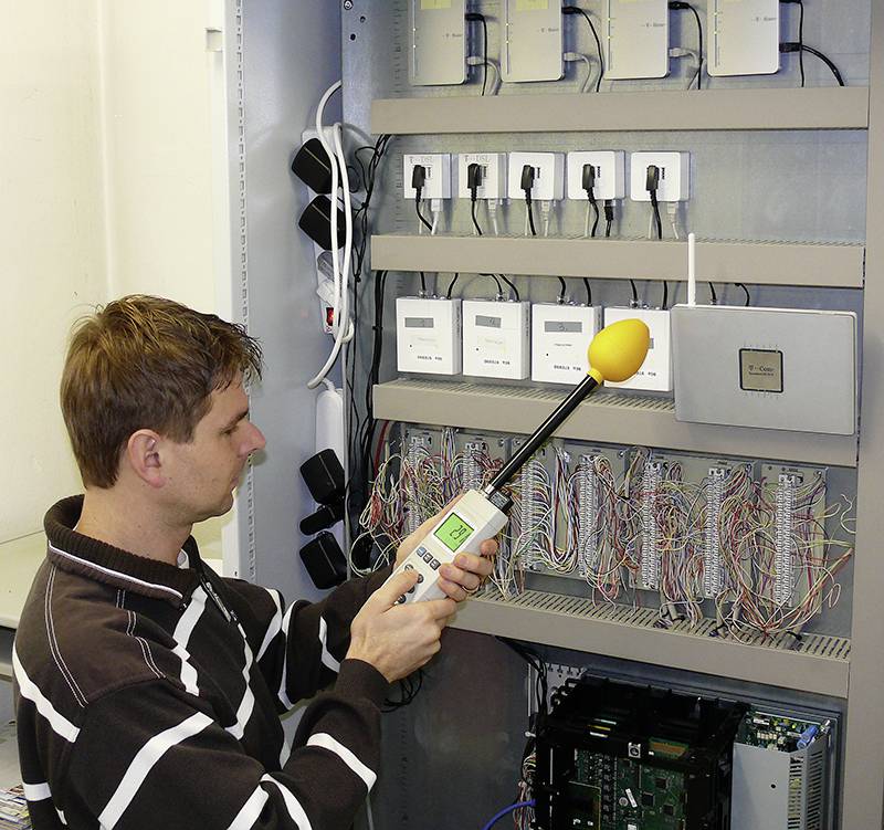 Ein Mann in gestreiftem Pullover misst mit einem Schallpegelmessgerät die Geräusche an einem offenen Schaltschrank voller Kabel und Geräte.