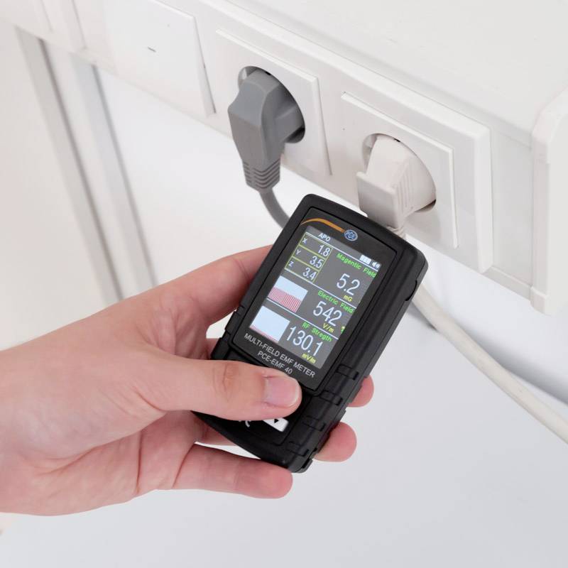 Eine Hand hält ein Leistungsüberwachungsgerät, das Daten von einer Steckdosenleiste misst. Anzeigen zeigen Volt, Ampere, Watt.