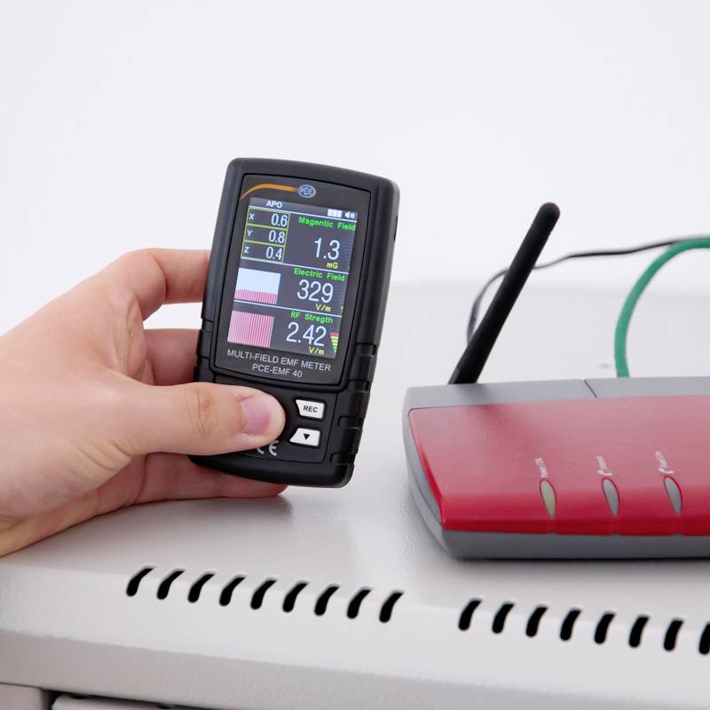 Eine Person hält ein EMF-Messgerät neben einen Router, um elektromagnetische Felder zu messen.