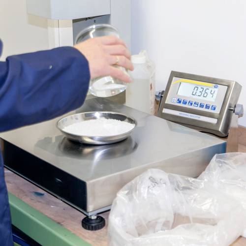 Eine Person hält ein Glasbehälter und schüttet Pulver auf eine Waage. Die Anzeige zeigt '0,364'. Wissenschaftliche Messung.