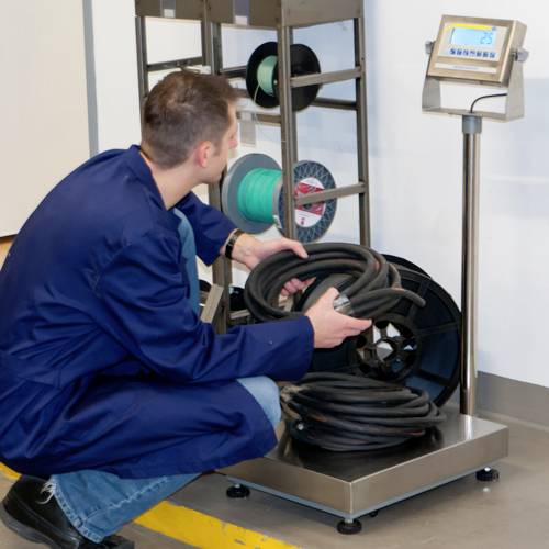 Ein Mann in blauer Arbeitskleidung kniet vor einer Waage und wiegt ein Kabel. Rechts neben ihm steht ein Display mit dem Gewicht.