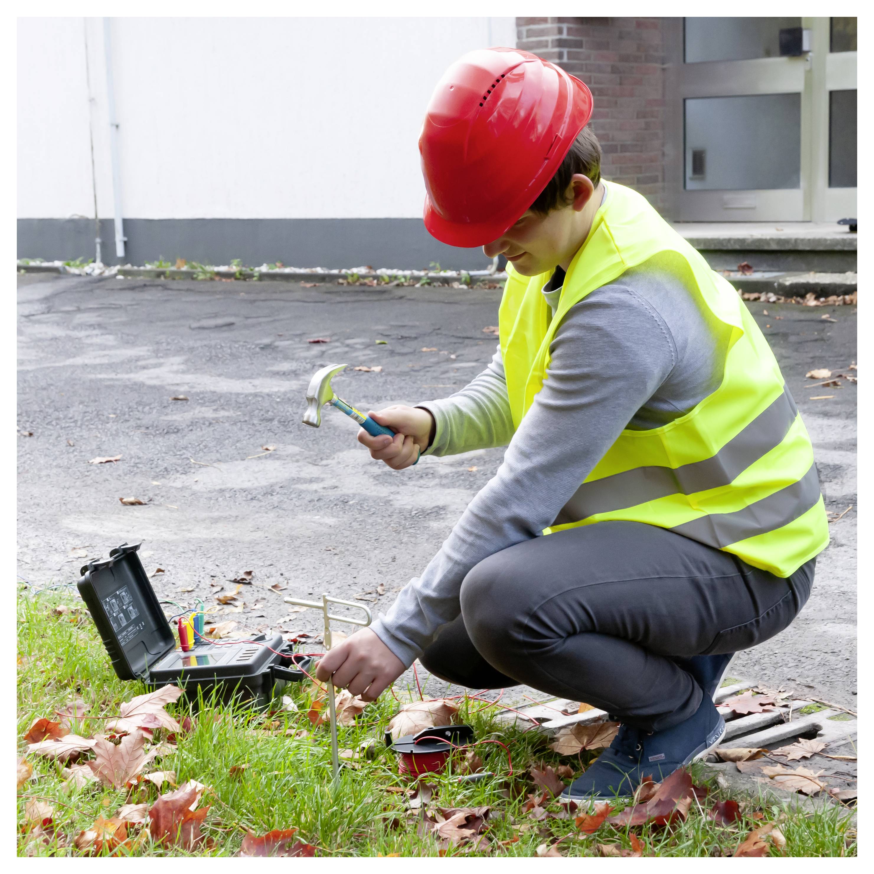 Arbeiter mit rotem Schutzhelm und reflektierender Weste kniet im Freien und verwendet Werkzeuge, um Verkabelung im Boden zu inspizieren oder zu reparieren.