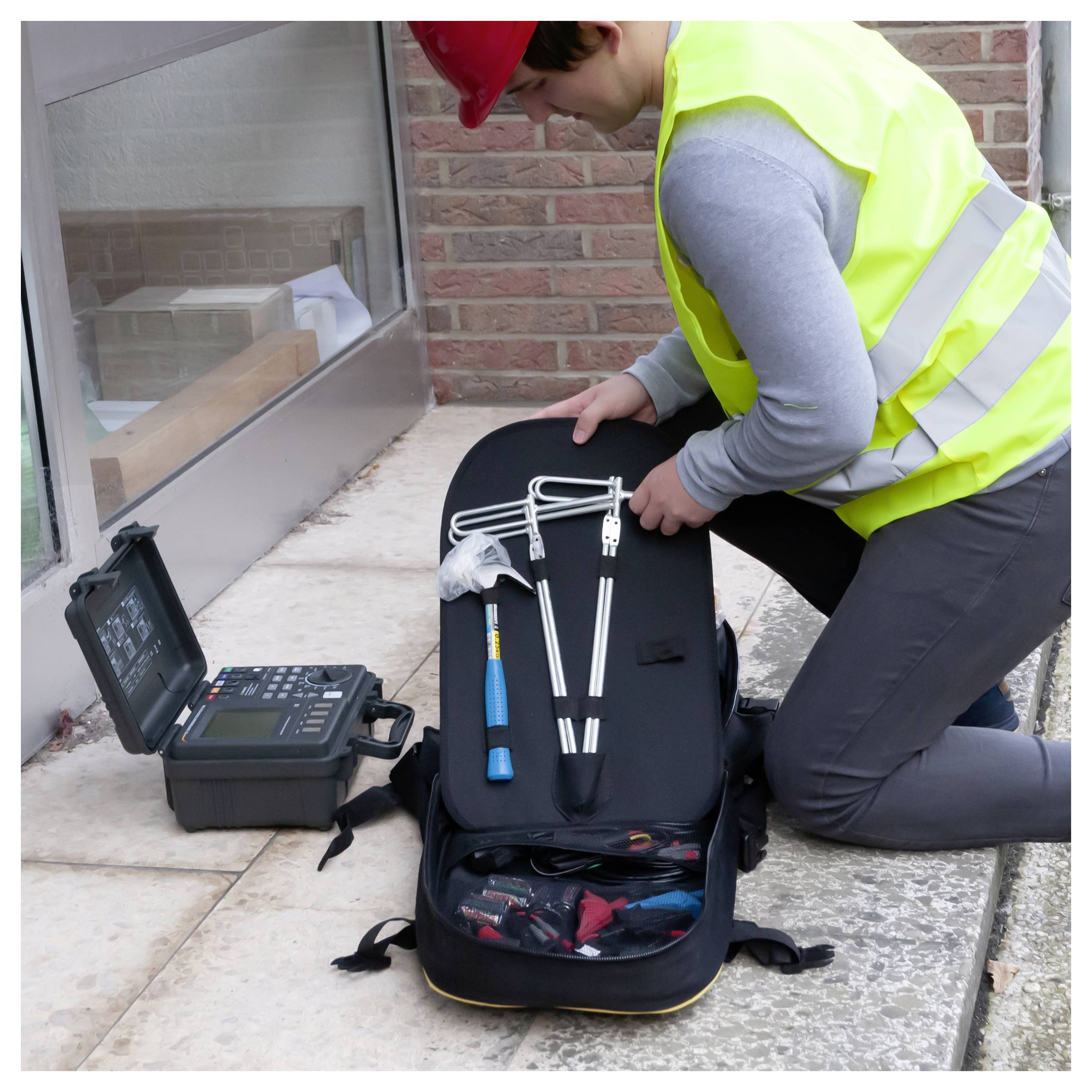 Techniker kniet mit einer offenen Werkzeugtasche und Inspektionsgeräten neben einem Fenster eines Gebäudes und bereitet sich auf die Arbeit vor.
