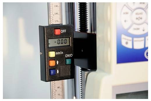 Digitales Messinstrument auf einer vertikalen Skala mit Anzeige 0.00 und mm/in-Steuerung, montiert auf einer Prüfmaschine.