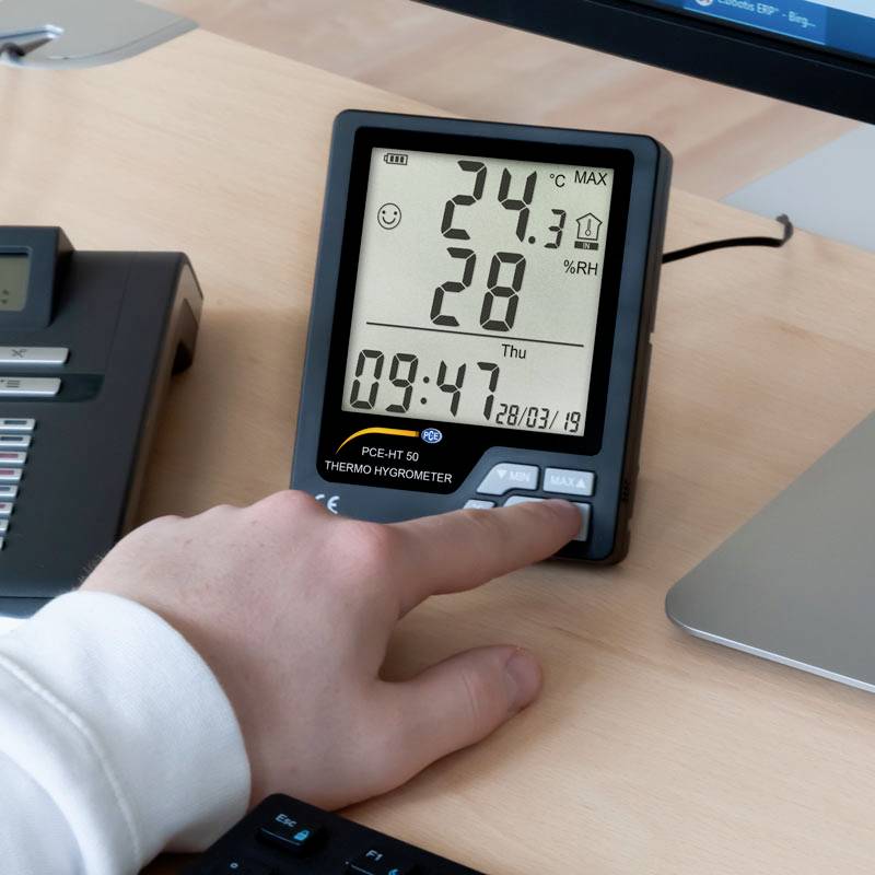 Eine Person bedient ein digitales Thermo-Hygrometer, das 24,1°C und 28% Luftfeuchtigkeit anzeigt. Ein Telefon und ein Monitor sind im Hintergrund sichtbar.