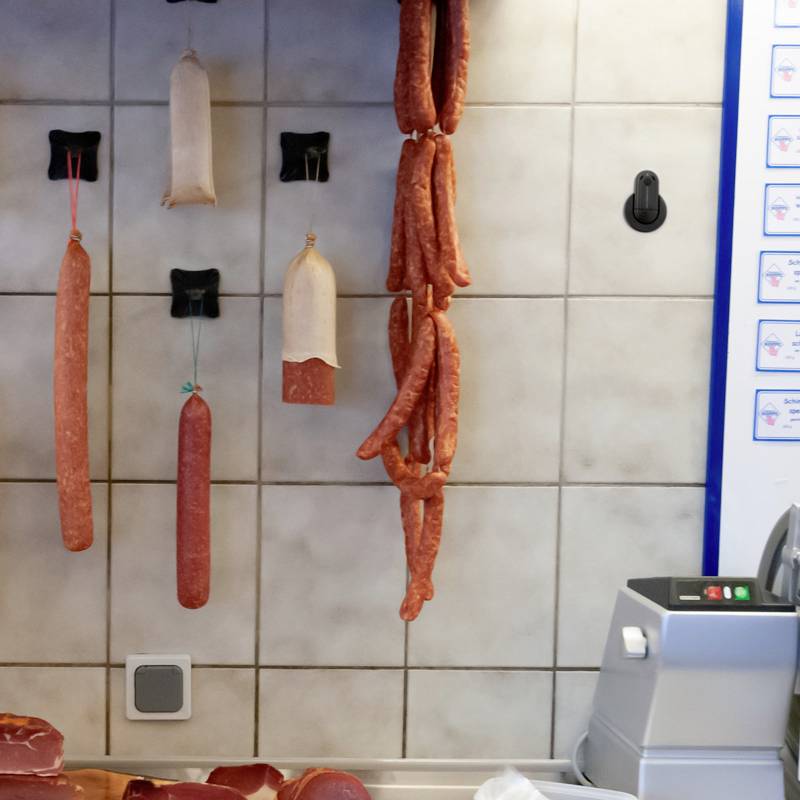 Verschiedene Wurstsorten hängen an Haken an einer gekachelten Wand in einem Fleischereifachgeschäft.