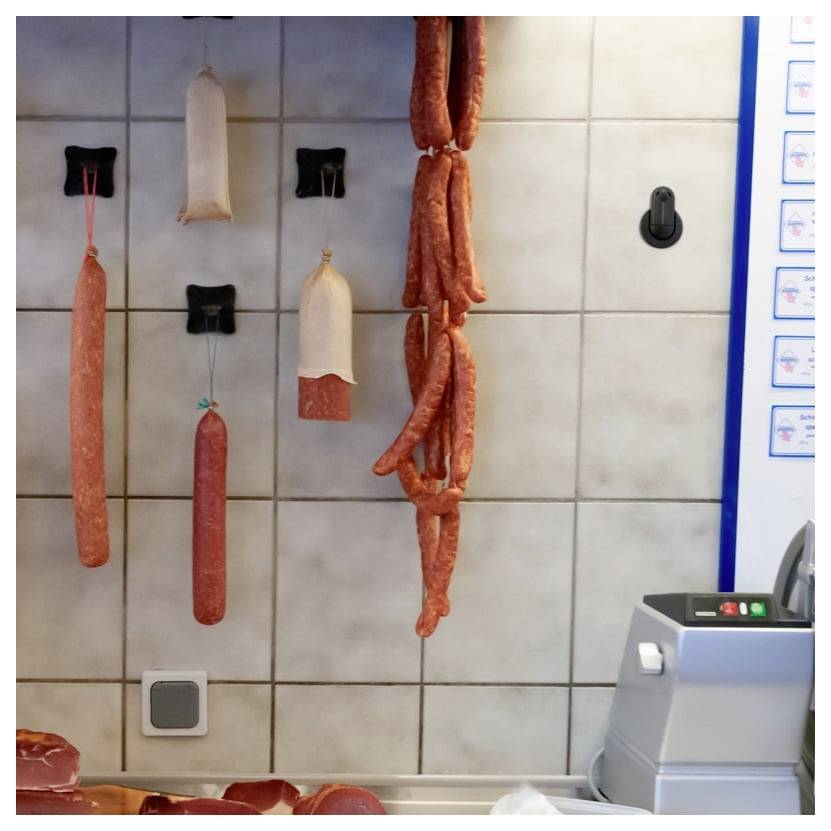 Mehrere Würste und Wurstwaren hängen an Wandhaken in einem gefliesten Metzgerei- oder Feinkostladen.