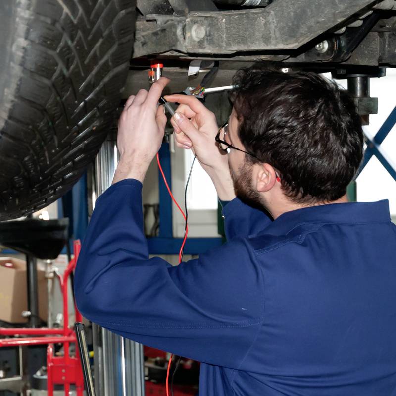 Ein Mechaniker arbeitet unter einem Auto auf einer Hebebühne, überprüft die Elektronik mit einem Diagnosegerät in einer Werkstatt.