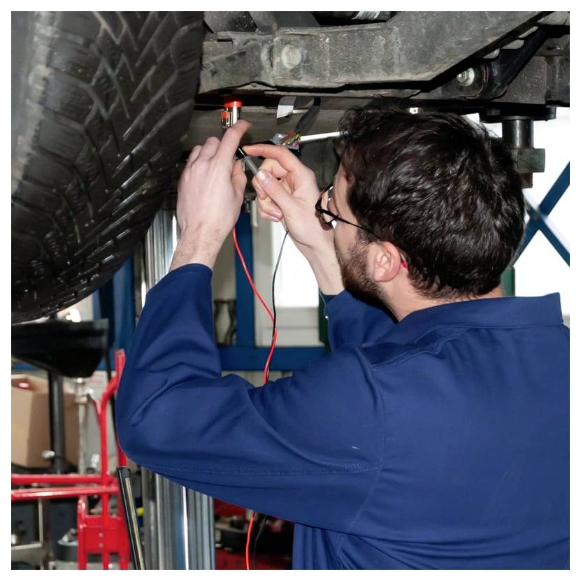 Mechaniker in blauem Overall verwendet ein Prüflicht und eine Sonde, um die Verkabelung unter einem auf einer Hebebühne angehobenen Fahrzeug zu überprüfen.