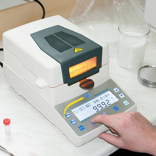Ein Laborgerät misst den Feuchtigkeitsgehalt eines weißen Granulats auf einer digitalen Anzeige. Eine Hand bedient das Gerät.