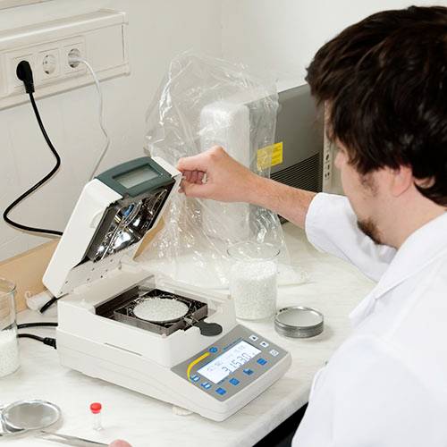 Person bedient ein Laborgerät zur Analyse von Proben. Ein geöffnetes Gerät mit Probenplatten ist zu sehen. Umgebung eines Labors sichtbar.