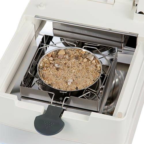 Eine Waage mit einem runden Behälter voller Sand und kleiner Steine zur Analyse.