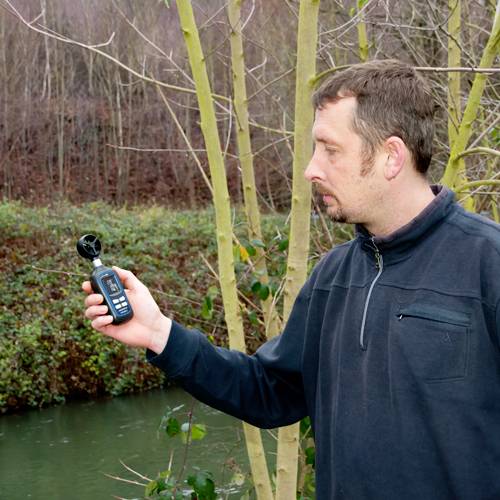 Ein Mann misst mit einem Handgerät die Windgeschwindigkeit im Freien in der Nähe eines Flusses, umgeben von kahlen Bäumen.