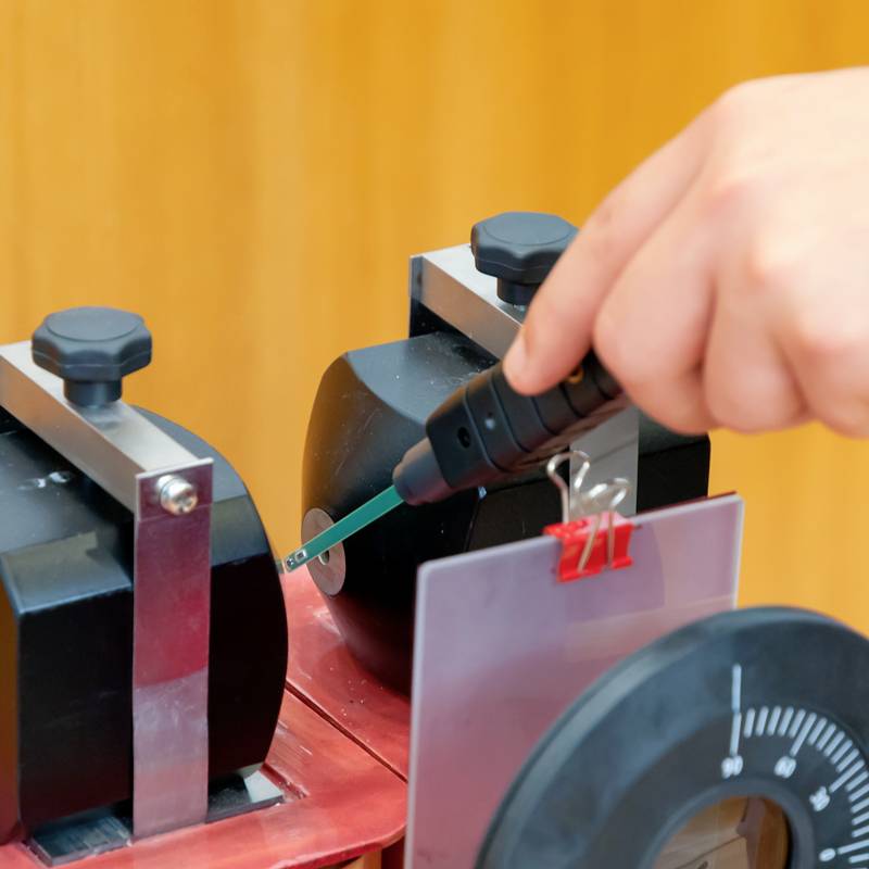 Eine Hand hält ein grünes Spektrometer vor ein Gerät zur Lichtmessung. Ein Zifferblatt ist im Vordergrund zu sehen.