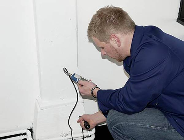 Ein Mann in Arbeitskleidung benutzt ein Messgerät an einer weißen Wand. Er misst möglicherweise Feuchtigkeit oder andere Parameter.