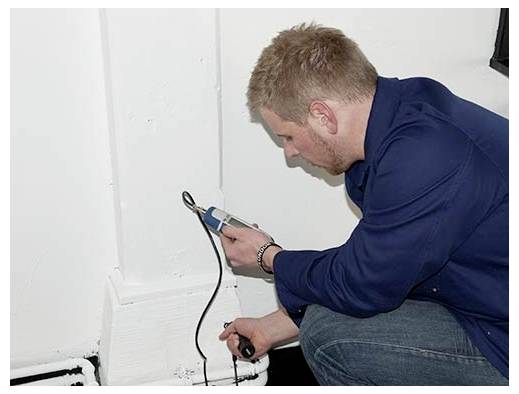 Ein Mann hockt sich an einer Wand nieder und inspiziert oder testet mit einem tragbaren Werkzeug eine Steckdose und ein Kabel in der Nähe des Bodens.