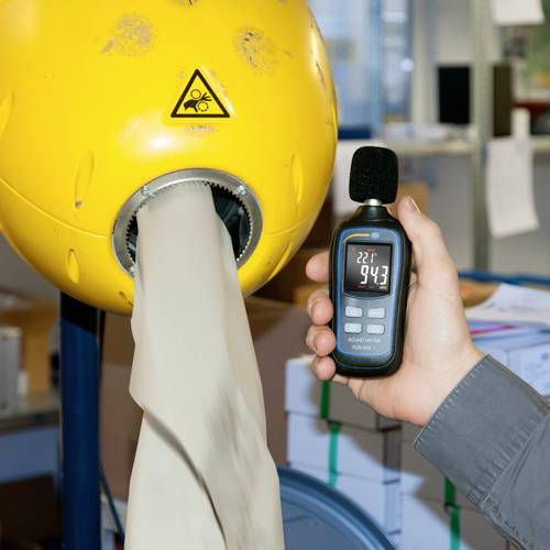 Eine Person hält ein Thermometer, das 22,7°C anzeigt, neben einer Maschine, die Mehl produziert. Ein Warnsymbol ist auf der Maschine.