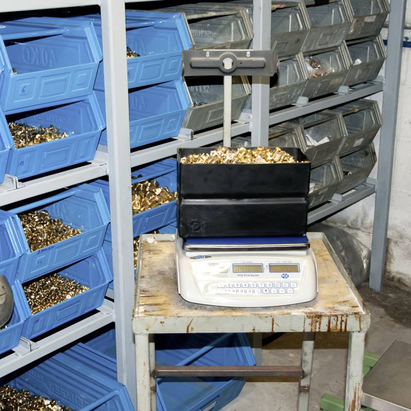 Metallteile in blauen Kisten auf Regalen. Vordergrund: Waage mit gefülltem Kunststoffbehälter. Arbeitet in einer Werkstatt oder Lagerumgebung.