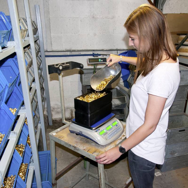 Eine Frau wiegt eine große Menge goldener Patronenhülsen auf einer digitalen Waage in einer industriellen Umgebung.