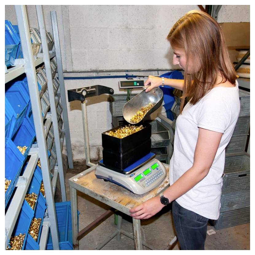 Eine Frau gießt goldfarben glänzende Münzen aus einer Schaufel in einen waagengefüllten Behälter in einem Lagerraum mit Regalen voller blauer Behälter.